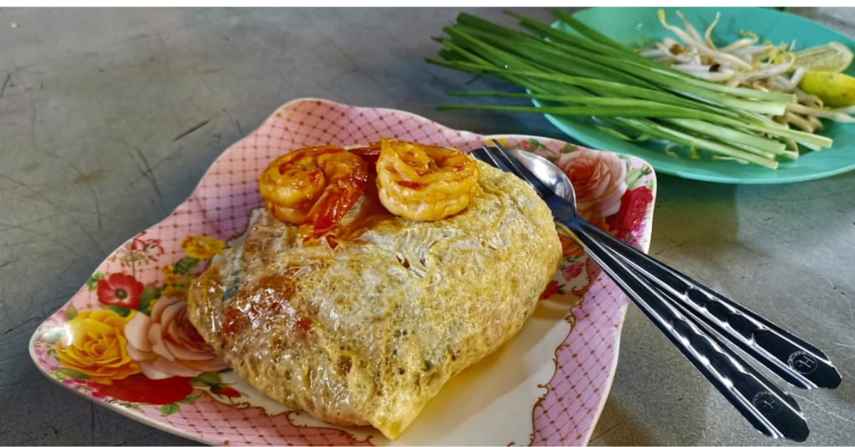 A pink plate with a serving of Pad Thai on it that is wrapped inside a layer of egg with two shrimp on top of it.