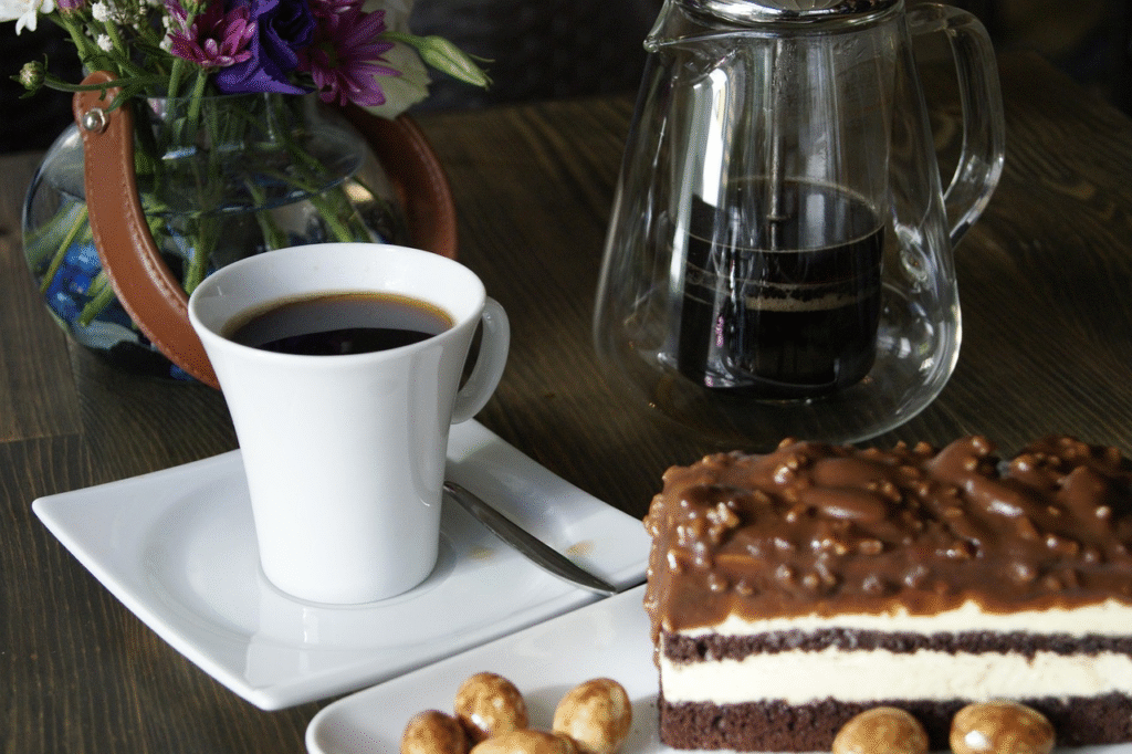A close-up of black coffee with a chocolate nut-covered cake on a dark wooden table, with a small vase of purple and white flowers in the background.

