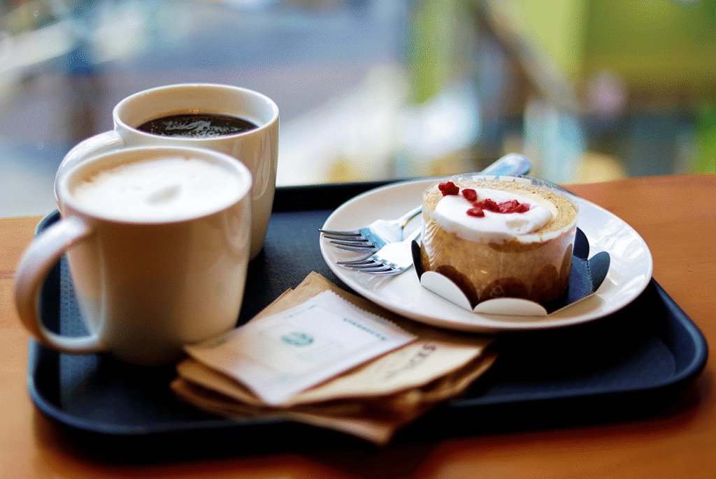 Two white mugs (black coffee and latte) with a slice of cream-topped Swiss roll on a black tray by a bright window.
