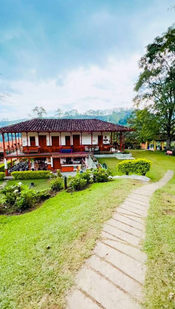 This image shows a traditional-style house with a red-tiled roof, surrounded by a lush green lawn and plants. In front of the house, a stone pathway leads upwards through the lawn. In the background, hills and trees are visible under a light blue sky.